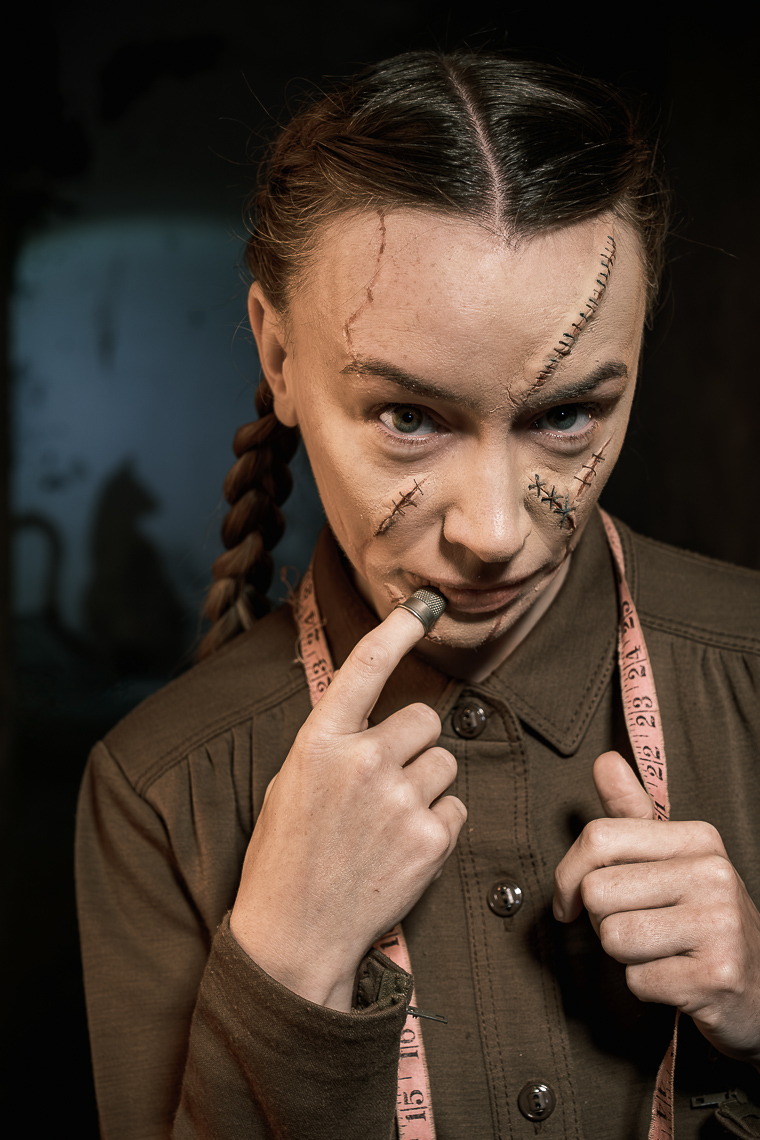 A promotional portrait photograph by a leading Cork-based theatre photographer Marcin Lewandowski, featuring actress Irene Kelleher. She is shown from the chest up, with dark braided hair pulled back, looking directly at the viewer with an intense expression. Her face is adorned with theatrical makeup designed to resemble prominent scars and stitches, particularly on her forehead and across her right cheek and nose. She wears a dark, possibly olive green, collared shirt with buttons, and a light pink measuring tape is draped around her neck. Her right hand is raised, with her index finger touching her lips, which are also painted with what appears to be a stitched effect. In the dark, blurred background to the left, a faint, dark silhouette resembling a cat's head with prominent ears is visible.