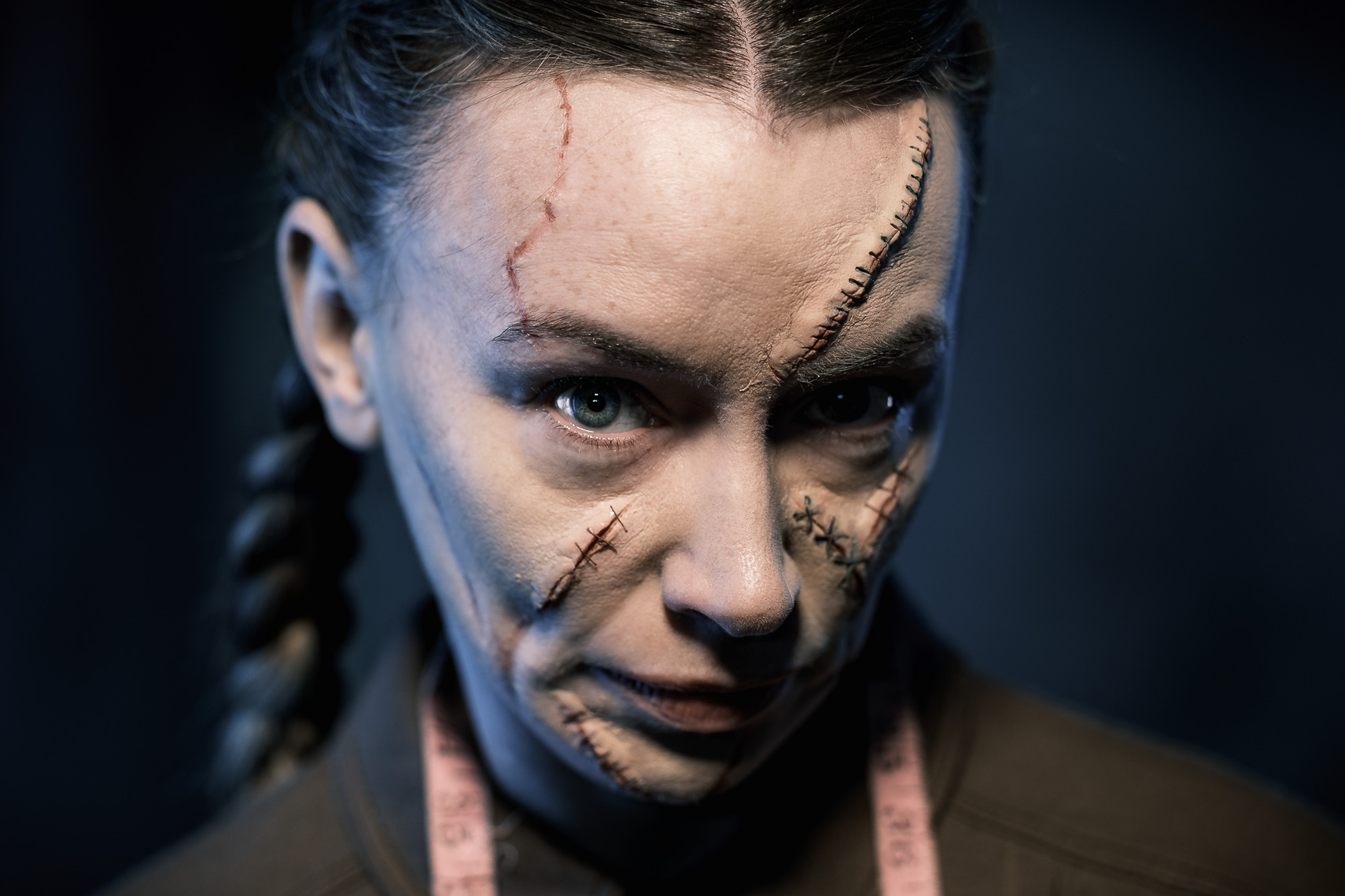 A promotional portrait photograph by a leading Cork-based theatre photographer, featuring actress Irene Kelleher for a theater play. She has braided dark hair and looks directly at the viewer. She has theatrical makeup on her face, designed to resemble scars and stitches. A prominent "scar" runs diagonally across her forehead, and another, with visible "stitches," covers a significant portion of her right cheek and nose. A light pink measuring tape is draped around her neck. The background is dark and out of focus.