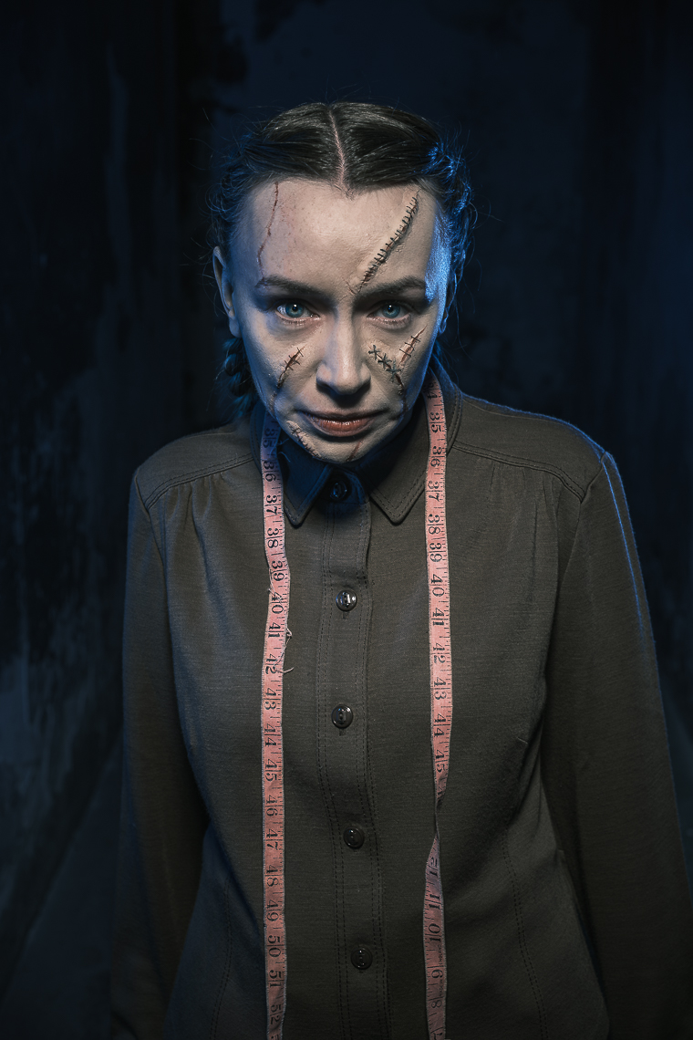 A promotional portrait photograph by leading Cork-based theatre photographer Marcin Lewandowski, featuring actress Irene Kelleher. She is shown from the torso up, looking directly at the viewer with a neutral expression. Her dark hair is pulled back into two braids, and her face is adorned with theatrical makeup designed to resemble prominent scars and stitches, particularly across her forehead and her right cheek and nose. She wears a dark, possibly olive green, collared shirt with buttons, and a light pink measuring tape is draped around her neck. The background is dark and indistinct, with subtle hints of a textured surface on the left and right.