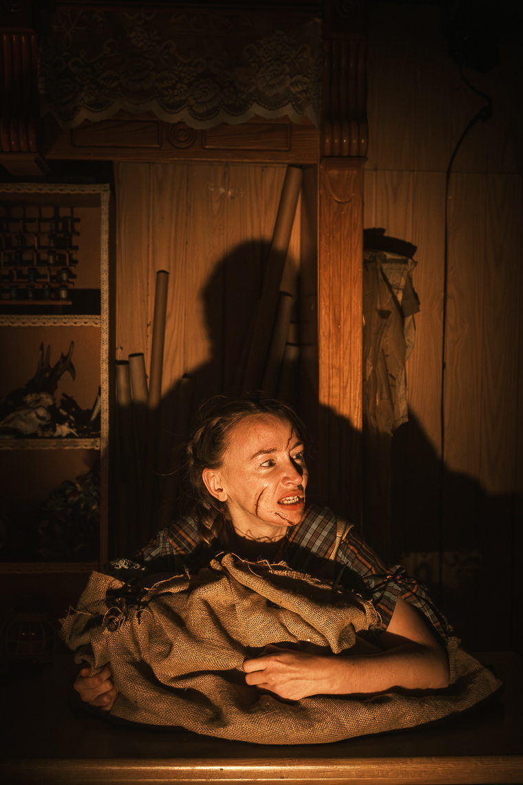 A dramatic production photograph by leading Cork-based theatre photographer Marcin Lewandowski, from Irene Kelleher's play "Stitch." The image captures a person, likely Irene Kelleher, in a distressed state. They are hunched forward, clutching a large, crumpled burlap sack. Their face is contorted in an expression of fear or anguish, with visible streaks of dirt or makeup on their cheeks and chin, and their eyes wide. They are wearing a plaid shirt. The setting appears to be an old, cluttered room with wooden paneling and shelves in the background. Warm, directional lighting casts deep shadows, highlighting the figure and creating an intense, atmospheric mood.