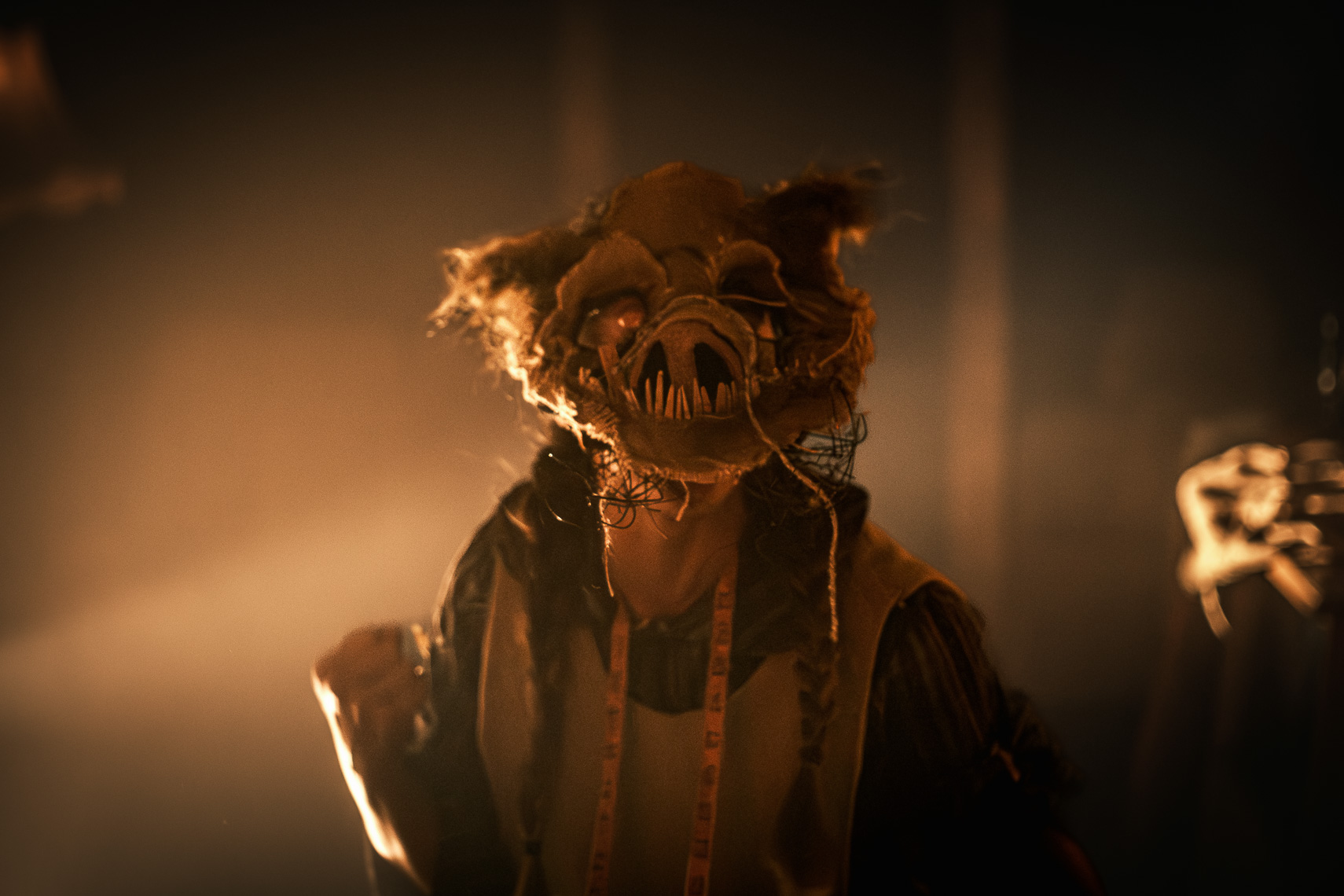 A striking production photograph by leading Cork-based theatre photographer Marcin Lewandowski, from Irene Kelleher's play "Stitch." The image features a person from the chest up, shrouded in a theatrical, dimly lit atmosphere. They are wearing a grotesque, monstrous mask resembling a pig's head, made from what appears to be burlap or rough fabric. The mask has sharp, prominent teeth, unsettling eyes that seem to glow, and dishevelled "hair" or straw-like elements. A small, dark spider prop is visible near the person's neck. They are wearing dark clothing, possibly a shirt with horizontal stripes, and a light-colored measuring tape is draped around their neck. The background is dark and hazy, with warm, theatrical lighting illuminating the figure from the front.
