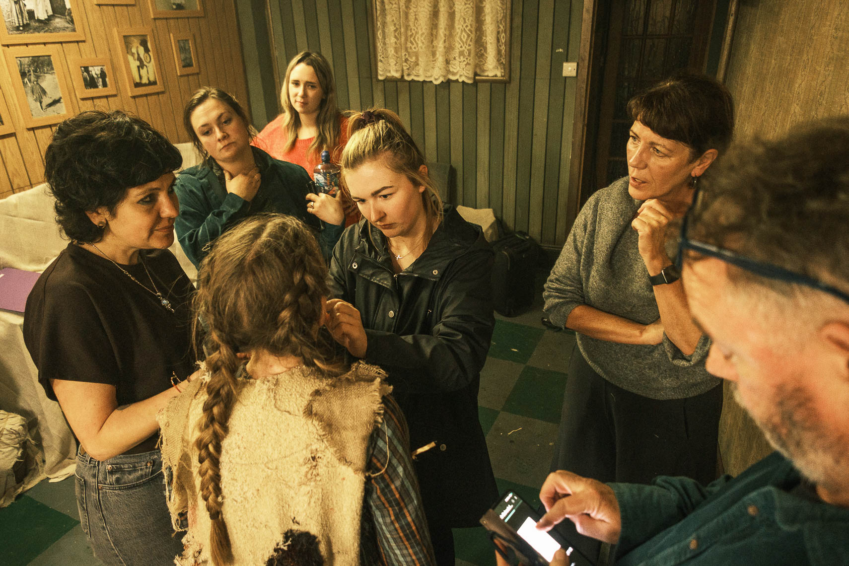 A behind-the-scenes production photograph by leading Cork-based theatre photographer Marcin Lewandowski, from Irene Kelleher's play "Stitch." The image shows a group of six individuals in what appears to be a backstage or dressing room setting, with wooden paneling and framed pictures on the wall. In the center, facing away from the camera, is a person with braided hair wearing a distressed, light-colored vest over a plaid shirt, likely actress Irene Kelleher in costume. Around this central figure, five other individuals are engaged in various tasks. One woman on the left, with dark, curly hair, is looking towards the costumed person. Another woman behind her, with dark hair and a teal top, is looking on. A woman in a black jacket is adjusting something on the costumed person's attire. To the right, a woman with short dark hair and a grey sweater is observing. In the foreground, partially visible on the far right, a person with a beard and glasses is looking down at a smartphone. The scene suggests a collaborative effort in preparing for the production, with a focus on costume or makeup details.