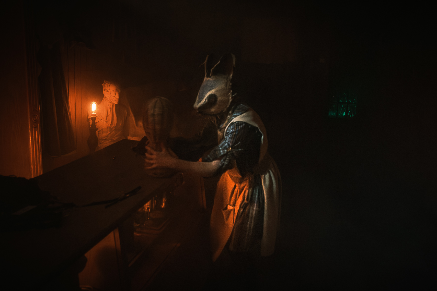 A darkly atmospheric production photograph by leading Cork-based theatre photographer Marcin Lewandowski, from Irene Kelleher's play "Stitch." The scene is set in a dimly lit, shadowy room. In the foreground, to the right, a figure wearing a disturbing, sack-like rabbit mask with long ears and possibly large teeth is hunched over a dark table. They are wearing a plaid shirt and a light-colored apron. Their hands appear to be manipulating an object on the table, which looks like a burlap head or doll. In the background, to the left, another figure, partially obscured and seemingly wearing a sack over their head, stands next to a window or opening, holding a single glowing light source, possibly a bare bulb or lantern, which casts an orange glow. The overall lighting creates a sense of suspense and unease.