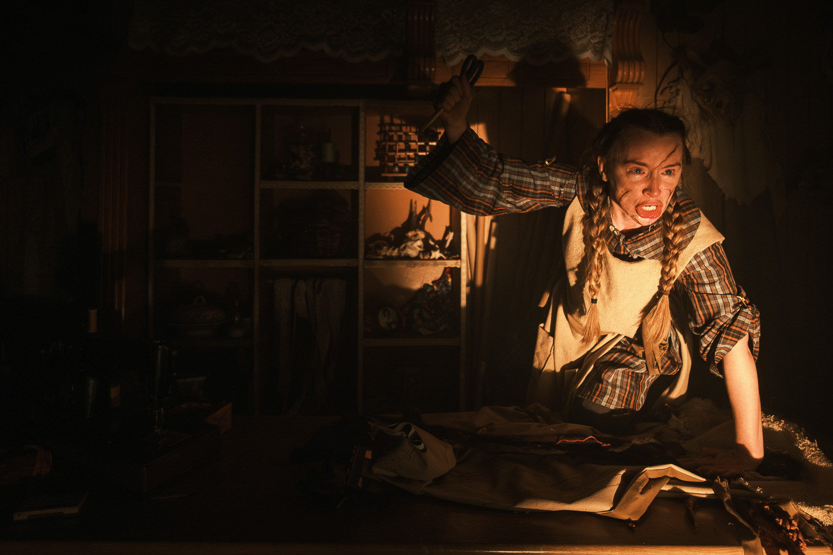 A tense production photograph by leading Cork-based theatre photographer Marcin Lewandowski, from Irene Kelleher's play "Stitch." The image shows a person, likely Irene Kelleher, in a dynamic and distressed pose. She is leaning forward, with her mouth open as if screaming or crying out, and her face is streaked with what appears to be dirt or makeup, and her eyes wide with emotion. Her hair is in two long braids. She is wearing a plaid shirt under a light-colored apron or vest. Her right arm is raised, holding an indistinct object, possibly a tool. In the foreground, a piece of fabric or clothing lies crumpled on a surface, possibly with faint reddish stains. The background features a dark wooden shelving unit with various items, creating a cluttered and confined atmosphere. The scene is lit dramatically, with strong shadows adding to the intensity.